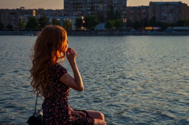 Nehir kenarında oturan rahat kızıl saçlı kadın. Güneş ışığı, yan görüş..
