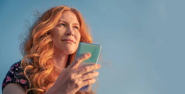 穿着夏装的漂亮的姜汁女士在天空前用智能手机 — 图库照片