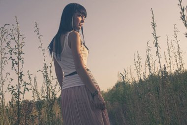 20 'li yaşlarda romantik elbiseli, yaz arkaplanındaki muhteşem genç bayan geriye bakıyor. Sarı kuru ot, loş gökyüzü. Güneşli bir akşam. Esmer model tek başına. Sakinlik ve denge..