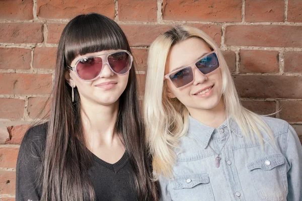 Zwei schöne, glückliche Mädchen in trendigen Sonnenbrillen vor städtischem Hintergrund oder roter Backsteinmauer. Junge Hipster. Außenporträt. — Stockfoto