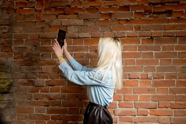 Une jeune blonde fait du selfie. Selfshot à l'extérieur contre le mur de briques rouges — Photo