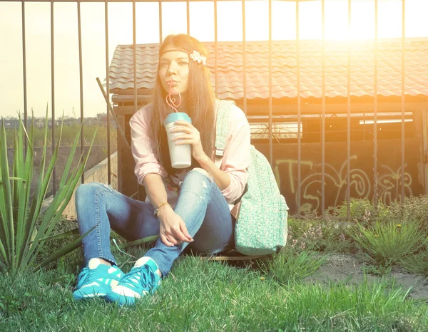 Feliz joven latina adolescente sosteniendo una taza de café para llevar sonriendo. Retrato de verano al aire libre de una hermosa adolescente sentada en la hierba cerca de la valla en el parque. Imagen llena de sol . — Foto de Stock