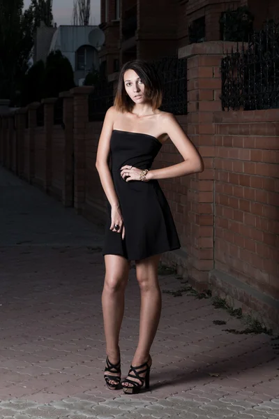 Linda mujer morena en vestido negro corto está posando contra la pared de ladrillo rojo —  Fotos de Stock