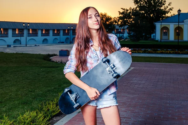 Ruiva menina está segurando skate em suas mãos ao pôr do sol — Fotografia de Stock