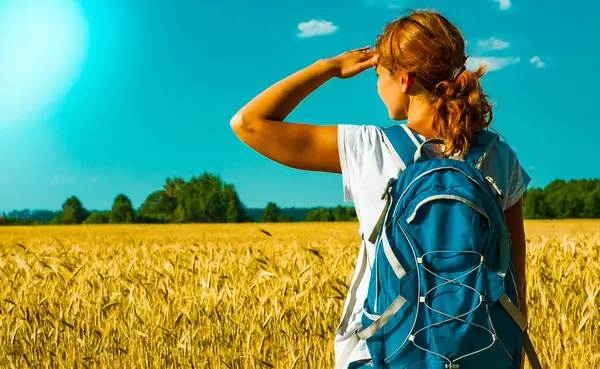 Young female tourist with backpack looking into the distance near a wheat field under the hot summer sky. back view — Stock Photo, Image