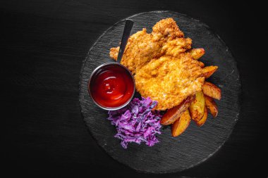 Ev yapımı Viyana şnitzeli patates ve salatayla siyah ahşap masa arkasında.