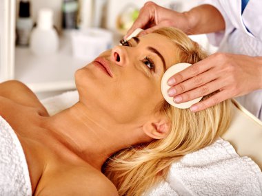 Woman middle-aged in spa salon and beautician cleanses face with sponge .