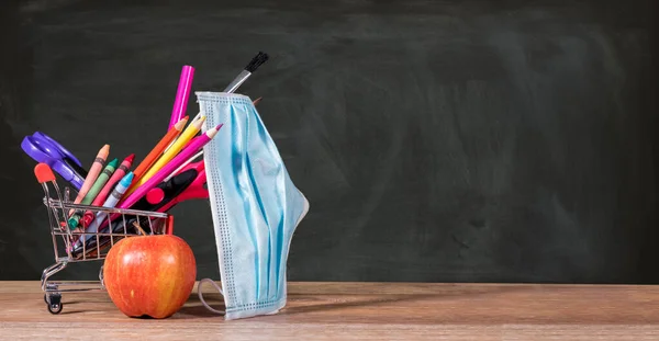 Back to school concept with pencils, crayons in shopping cart with apple with face mask — Stock Photo, Image