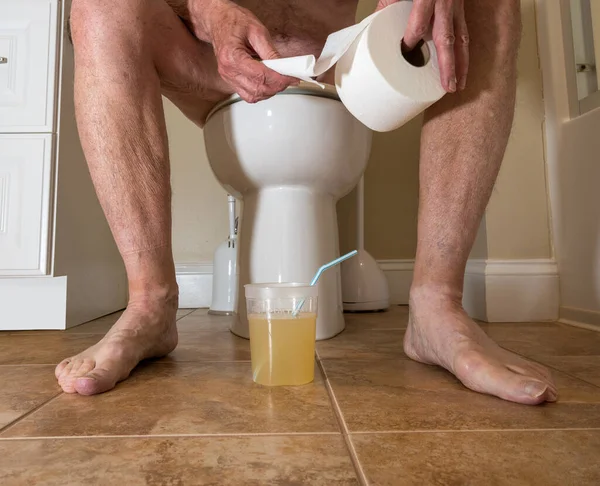Hombre adulto mayor sentado en el inodoro con rollo de papel y preparación de colonoscopia en el baño — Foto de Stock