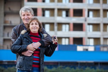 Mutlu gülümseyen beyaz çift ellerinde ev anahtarıyla dışarıda kucaklaşıyorlar.