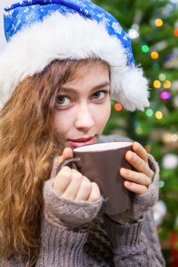 Noel Baba şapkalı genç bir kadın fincandan kahve içiyor ve kameraya bakıyor.