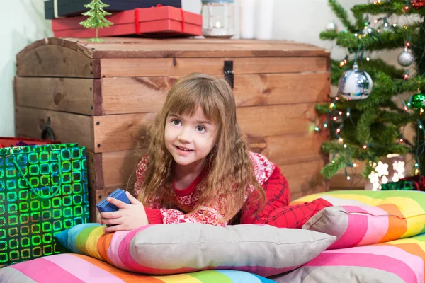 クリスマスツリーの近くの色の枕に横たわって 手にギフトボックスを保持就学前の女の子 — ストック写真