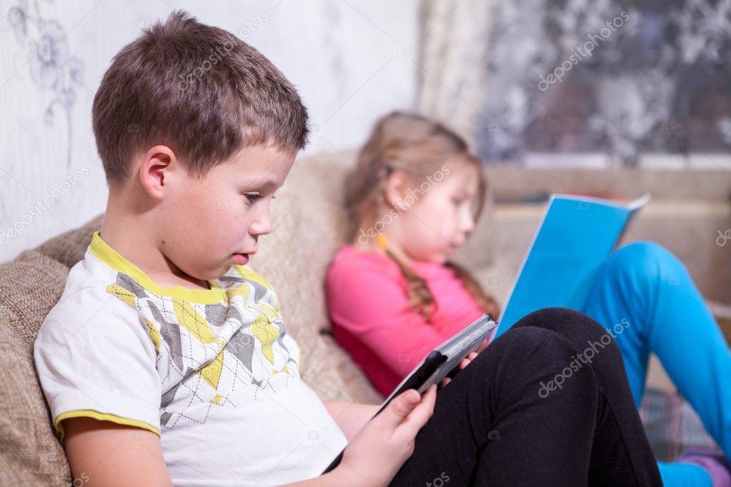 Brother sitting with electronic tablet in hands, sister reading a book on the couch in the room
