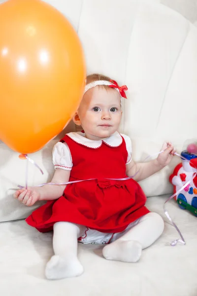 小さな赤ちゃん女の子で赤いドレス座っていますおもちゃとともにホワイトソファ — ストック写真
