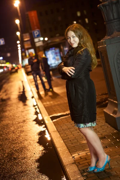 Jolie femme en manteau noir et talons hauts debout seul sur la route dans la ville de nuit — Photo