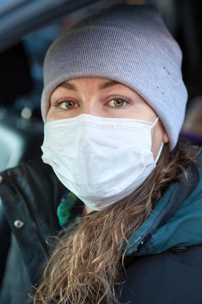 Curly hair Caucasian woman in a safety mask sitting inside her car, female wearing hat and coat