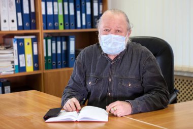 Kafkasyalı kıdemli iş adamı maske takıyor. Ofis odasında Covid-19 gribi yüzünden masada oturuyor.