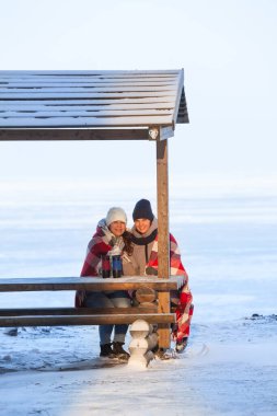 Ahşap bankta oturmuş, ısınmak için termo bardaklardan sıcak içecekler içen kırmızı eşarpla sarılan iki donmuş kız arkadaş.