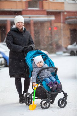 Genç anne, yeni yürümeye başlamış çocuğuyla birlikte bebek arabasında oturuyor, kışın açık havada boy portresi çiziyor.