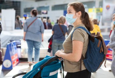 Uluslararası havaalanında bavulunu bekleyen çocuklu beyaz bir anne. Tıbbi maske takan bir kadın.