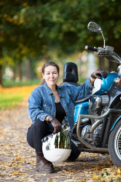 Female motorcycle rider with white crash helmet in hands sitting near her blue bike