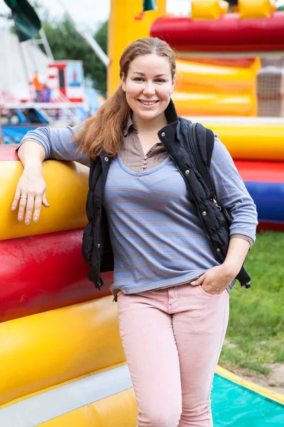 Toothy smiling woman portrait, wearing pink trousers and waistcoat, stands leaning to inflatable jump castle, outdoors portrait