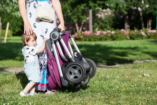 Mother calm down her toddler daughter while standing with pushchair in other hand, summer green lawn, copy space