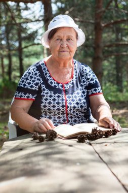 Yaşlı kadın kitap okuyor. Evergreen ormanındaki ahşap masada oturuyor ve kameraya bakıyor.