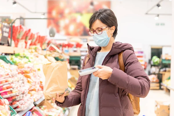 Woman doing groceries shopping at supermarket and wearing a face mask - Young woman buying fruit during coronavirus pandemic wearing protective mask - lifestyle and health