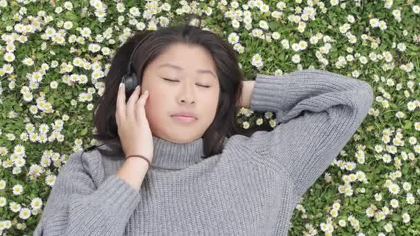Krásná mladá žena odpočívající v parku na jaře - Mladá asijská Číňanka ležící na trávě, poslouchající hudbu a užívající si čas v parku za slunečného dne v Londýně — Stock video