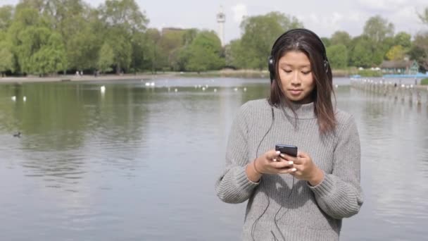 Bella giovane donna che si rilassa al parco e ascolta musica - Giovane donna cinese asiatica che si diverte al parco in una giornata di sole a Londra e utilizza il suo dispositivo intelligente per i social media — Video Stock