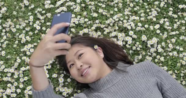 행복 한 동양인 소녀가 머리 사이에 데이지를 물고 셀카를 하고 정원을 뒤덮고 있었다 - 젊은 중국 여인 이 풀 위에 누워, 런던의 화창 한 날 공원에서 즐겁고 즐거운 시간을 보내고 있었다 — 비디오