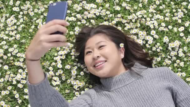 Chica asiática feliz tomando una selfie con una margarita entre el pelo y las margaritas jardín cubierto - Joven mujer china tumbada en la hierba, divertirse y disfrutar del tiempo en el parque en un día soleado en Londres — Vídeos de Stock