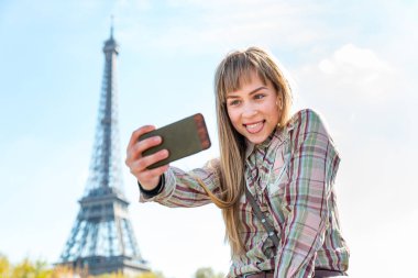 Paris 'te arka planda Eyfel Kulesi ile fotoğraf çeken mutlu bir kız sosyal medyada fotoğraf paylaşmak için akıllı telefonunu kullanan gülümseyen genç bir kadın - seyahat, teknoloji ve yaşam tarzı kavramları