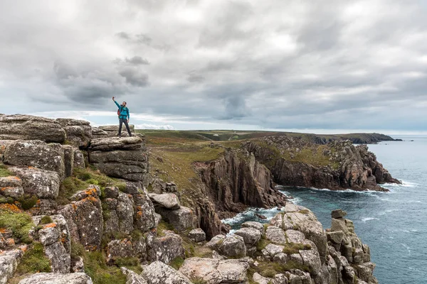 Man taking a selfie on a rock cliff enjoying the view - Young man with backpack and hiking clothes on the cliffs over seashore in Cornwall, UK - Travel, nature and wanderlust concepts.