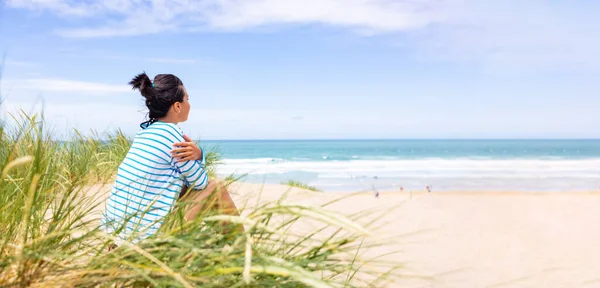 コーンウォールの海辺でビーチと波を見ている女性 晴れた日に海を背景に砂丘に座っている女性のリアサイドビュー 旅行と幸福の概念 — ストック写真