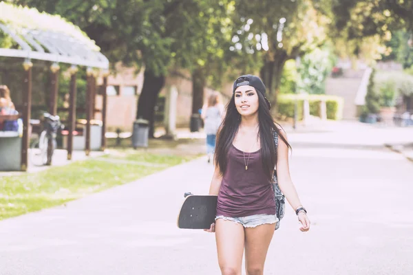 Hermosa chica caminando en el parque sosteniendo un monopatín . — Foto de Stock