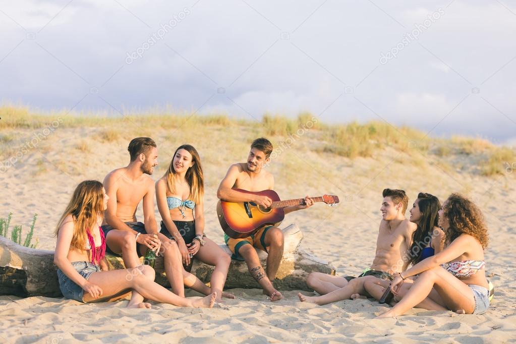 Group of friends playing guitar and singing on the beach at sunset. There are four girls and three boys, some are sitting on a log, some on the sand.