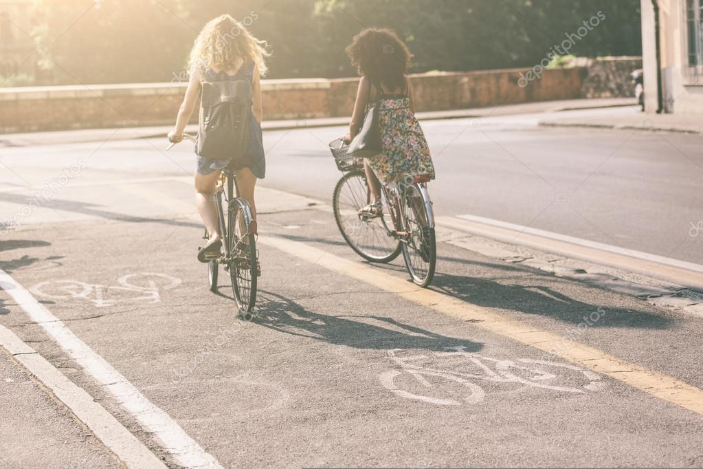 Couple Of Female Friends Riding Bikes On The Street Focus Bike Icon They Are Two Women Wearing Summer Clothes Couple Of Female Friends Riding Bikes On The Street Focus Bike Icon They Are Two Women Wearing Summer Clothes