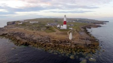 İngiltere 'de alacakaranlıkta uçurum manzaralı deniz feneri - Portland Adası' nın kenarında beyaz ve kırmızı bir deniz fenerinin insansız hava aracı videosu - seyahat ve mimari kavramları