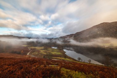 Vadi ve göl sisle kaplıydı, sonbahar yaprakları ve dağlarla bulutlu bir gökyüzü altında, sakin bir manzara yaratıyordu.