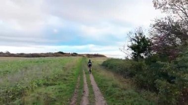 Kırsal kesimde manzaralı toprak bir patika boyunca dağ bisikleti süren aktif bir adam. Yeşil tarlaları ve uzaktaki denizi olan güzel bir manzara.