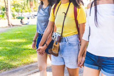 Genç bayan arkadaşlar el ele tutuşuyorlar, parkta birlikte yürüyorlar. Günlük yaz kıyafetleri giyip eski model bir kamera taşıyorlar.