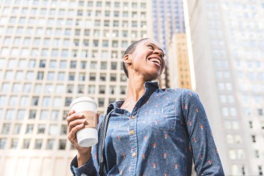 Kahve fincanı tutan mutlu siyah kadın, ofis binalarıyla dolu bir şehirde yukarı bakıyor ve gülümsüyor.