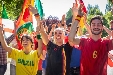 Takım forması ve bileklik takan, bayrak sallayan ve bir spor müsabakasında yüksek sesle tezahürat yapan çok ırklı gençler.