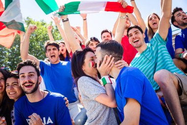 Bir grup heyecanlı İtalyan futbol fanatiği tezahürat yapıyor, kutlama yapıyor ve bayraklarla öpüşüp yüzlerini boyuyor.