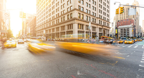 Busy road intersection in Manhattan, New York, at sunset. There are some blurred yellow cabs on foreground, and buildings, people and cars on background. Long exposure shot. Travel and city life.