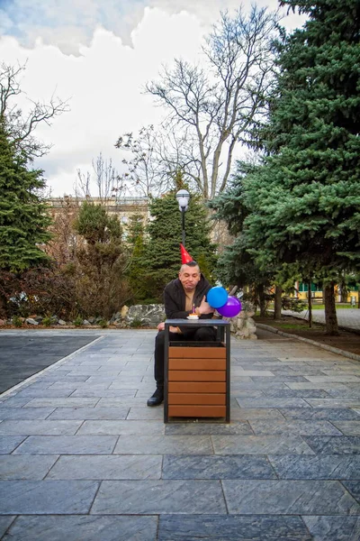 A sad birthday boy wearing a homemade party cap with balloons and cake with a candle celebrates his birthday alone in an empty park during an epidemic