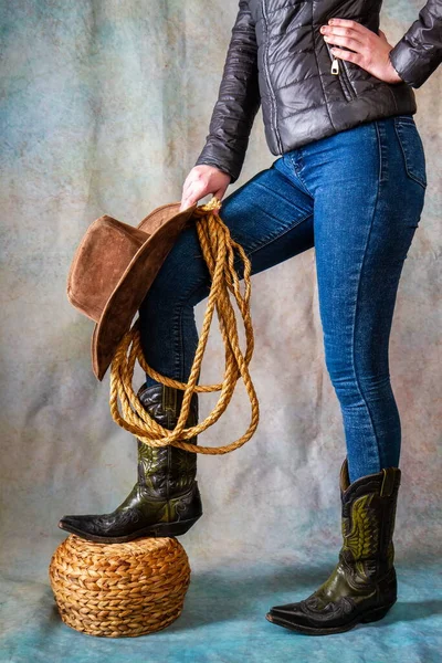 Female legs in leather cowboy boots and blue jeans with wide-brimmed hat and lasso on grunge background