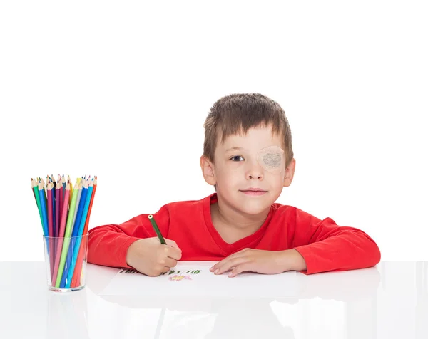 П'ятирічний хлопчик сидить за білим столом і малює олівці, ліве око застрягає гіпсом — стокове фото